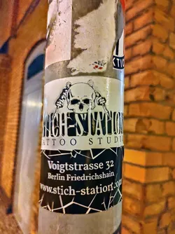 Street sticker Sticker found on a pole in Berlin, advertising Stich Station Tattoo Studio located on Voigtstrasse 32 in Friedrichshain. The sticker features a skull graphic and geometric design. 