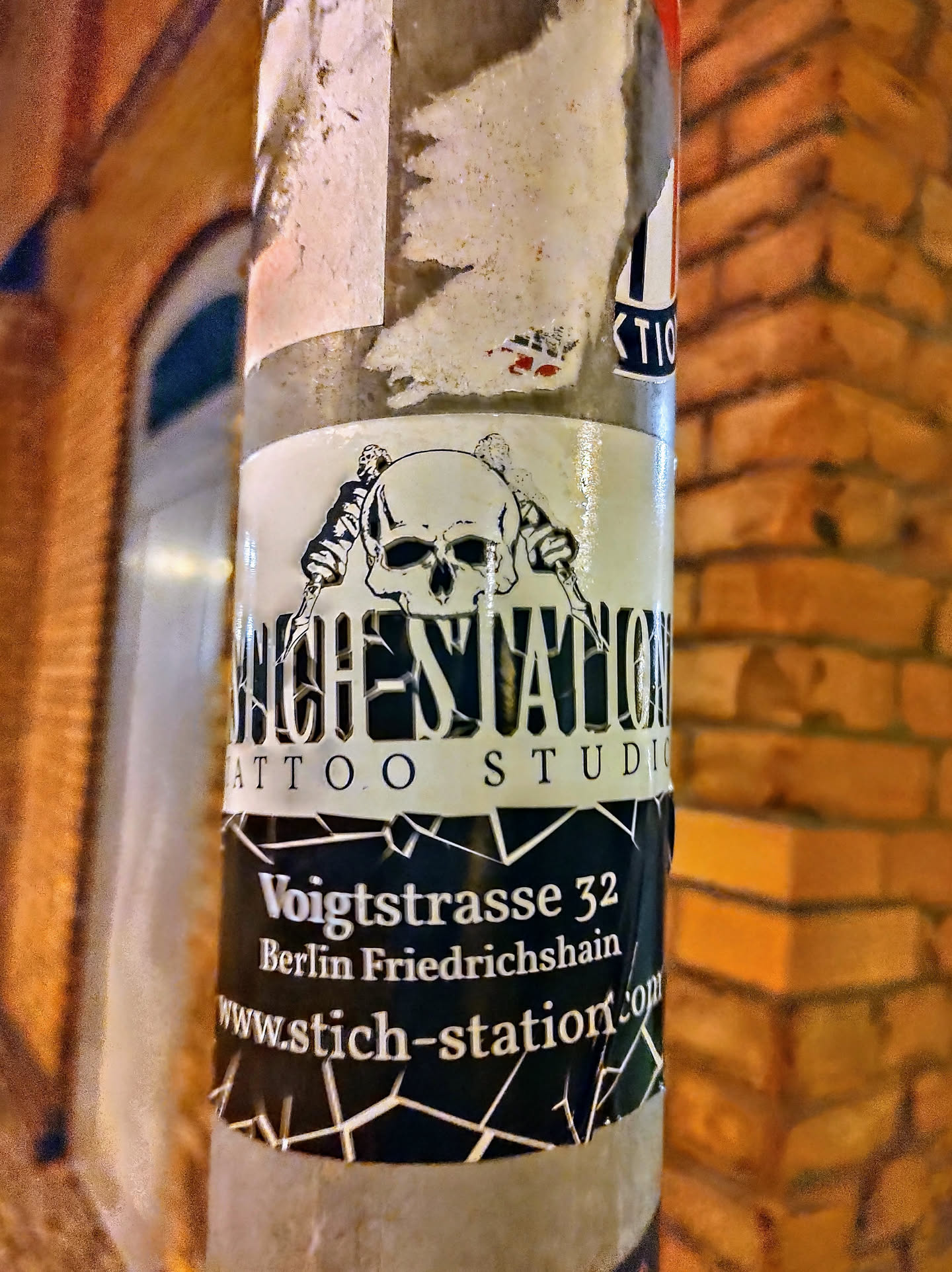 Sticker found on a pole in Berlin, advertising Stich Station Tattoo Studio located on Voigtstrasse 32 in Friedrichshain. The sticker features a skull graphic and geometric design. 