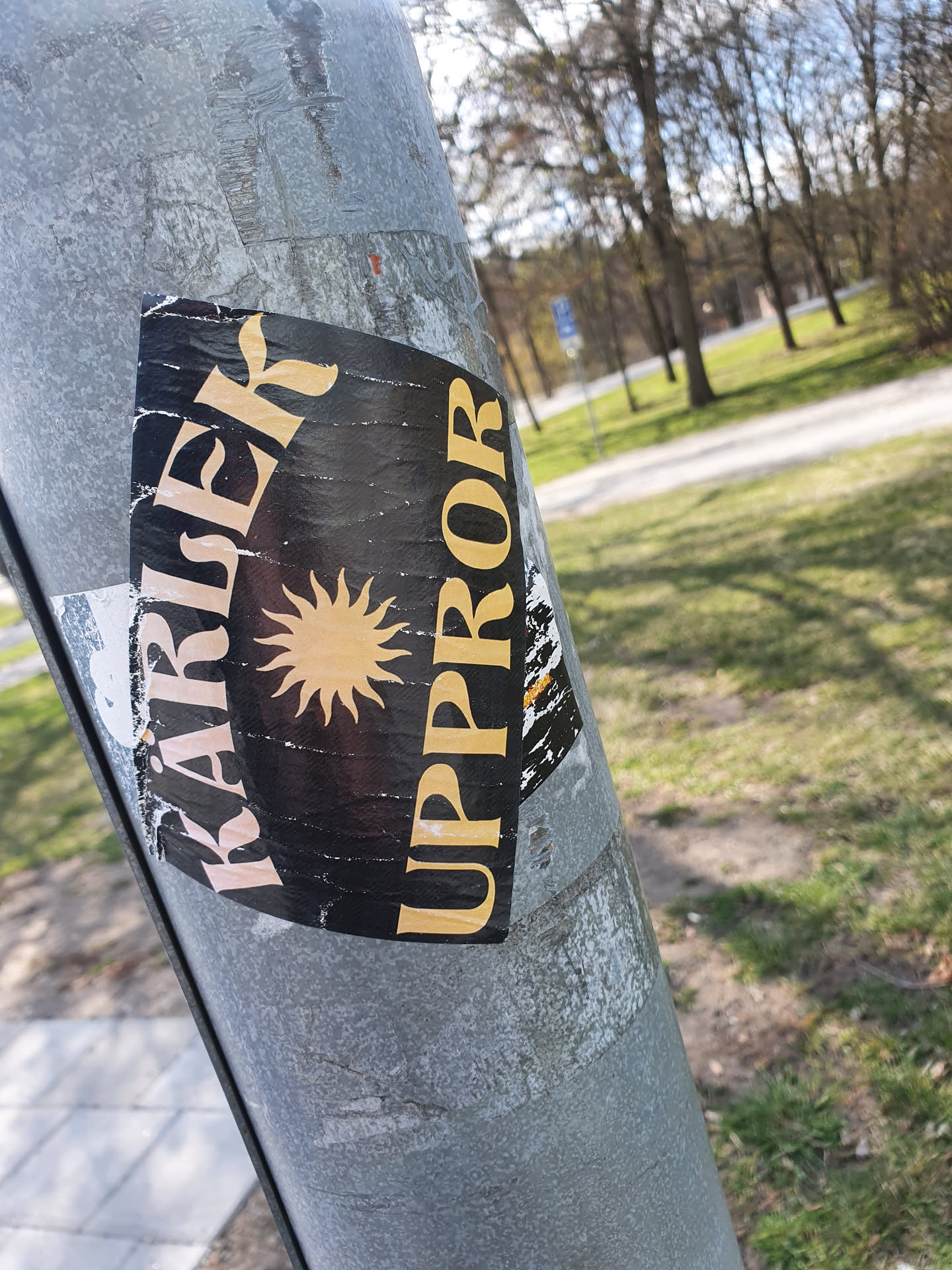 Close-up of a worn sticker affixed to a metal pole. The sticker is primarily black with yellow/gold text that reads "KARISIER UPPPROR." A stylized sun is centered on the sticker. The background shows a blurred park-like setting with trees and grass.