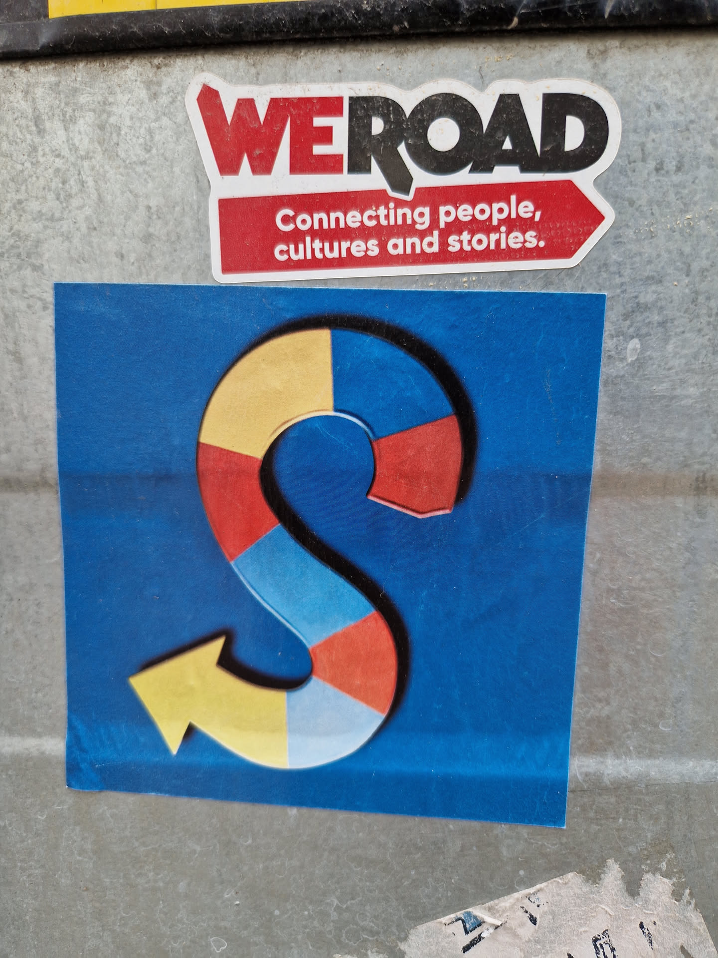 A sticker featuring a colorful arrow shaped as an S on a blue background. The upper sticker says: WE ROAD Connecting people, cultures and stories. The design suggests a journey, connection between cultures, and sharing of stories.