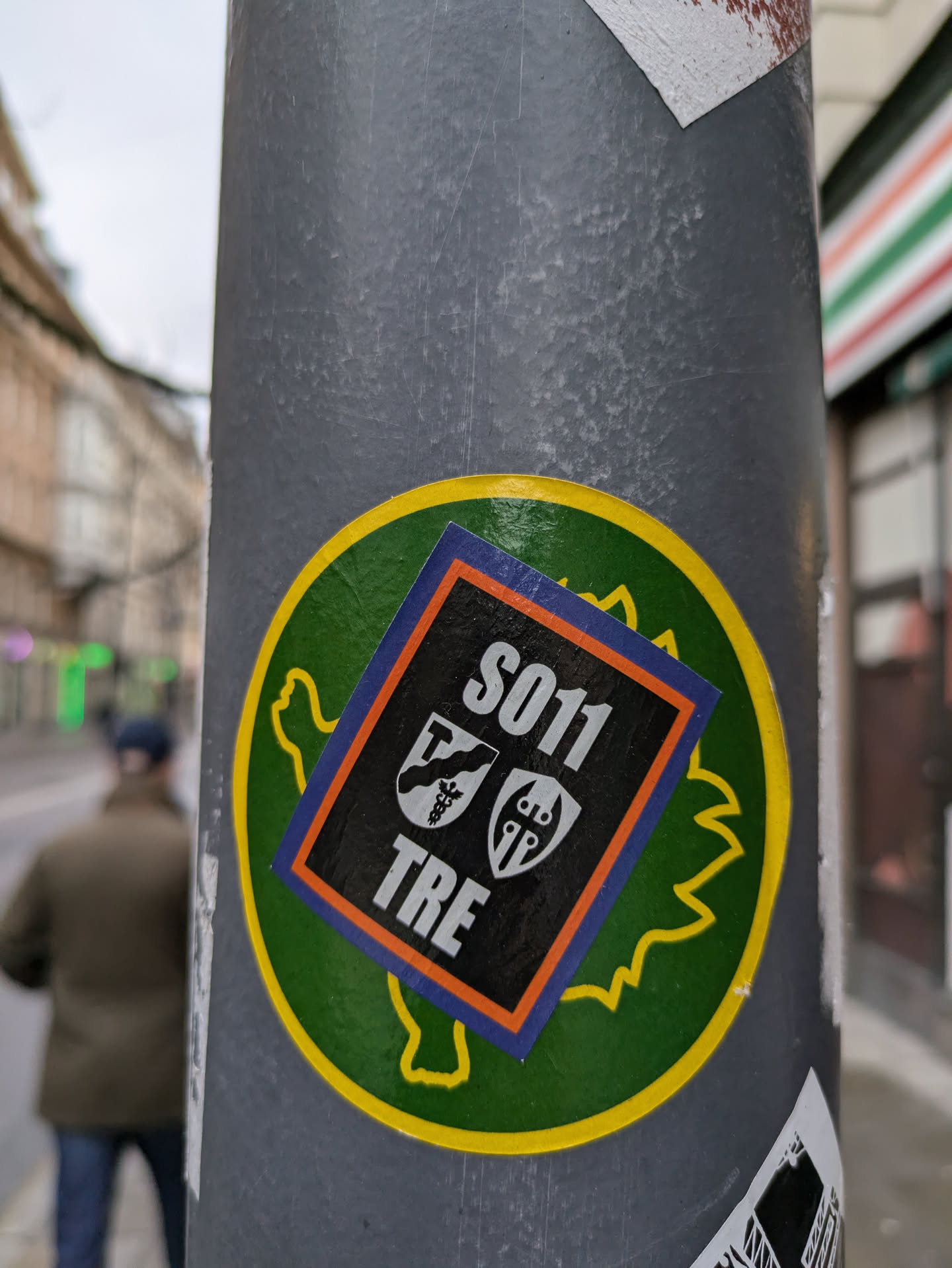 A black square sticker with the text S011 and TRE, along with two crest-like emblems, affixed to a larger round green sticker shaped like a stylized leaf or creature. The larger sticker is attached to a grey pole.