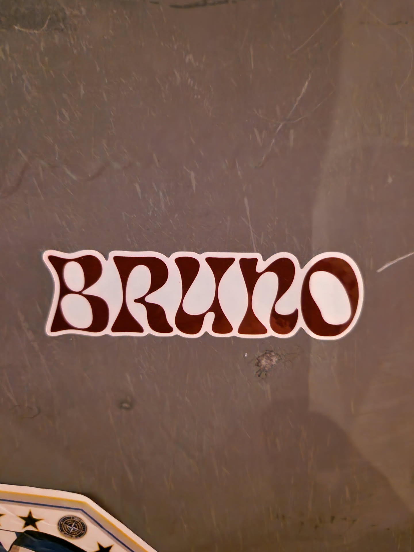 A maroon and white sticker with the name Bruno written in a retro, groovy font. The sticker is applied to a gray surface.