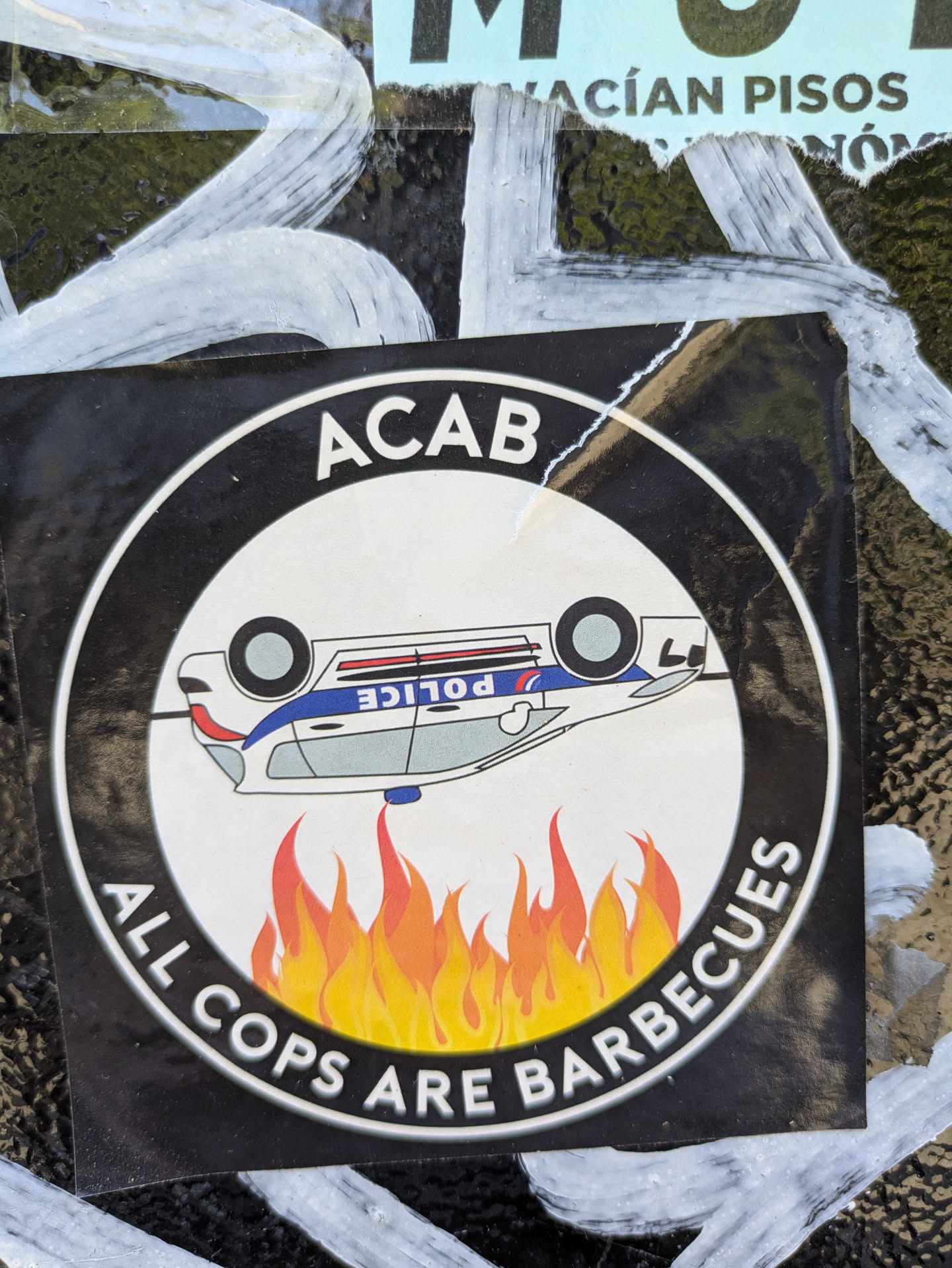 The sticker shows a black and white circular design. The text "ACAB" is at the top, and "ALL COPS ARE BARBECUES" is at the bottom. In the center, there is a cartoon of an upside down police car above flames.
