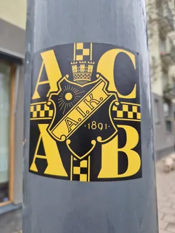 A close-up shot of a yellow and black sticker affixed to a gray pole. The sticker features a crest with the letters A.I.K. and the year 1891.  The letters A and B flank the crest, with a checkered pattern on either side.