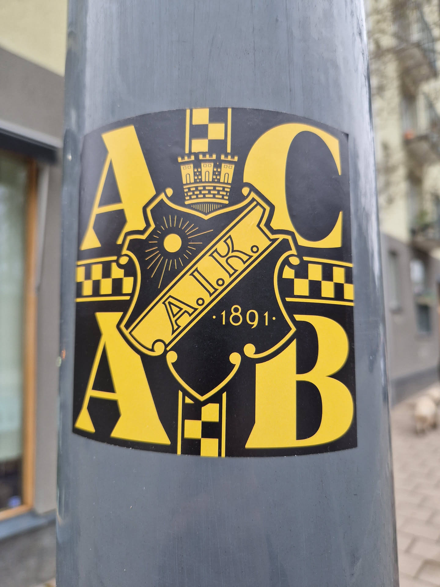 A close-up shot of a yellow and black sticker affixed to a gray pole. The sticker features a crest with the letters A.I.K. and the year 1891.  The letters A and B flank the crest, with a checkered pattern on either side.