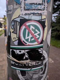 A sticker with a crossed-out logo, possibly an anti-fascist symbol. It is placed on a pole among various other stickers.