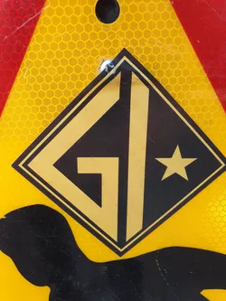Close-up of a diamond-shaped sticker with the letters "GI" in gold on a black background, and a gold star in the bottom right corner. The sticker is affixed to a yellow surface with a honeycomb pattern.