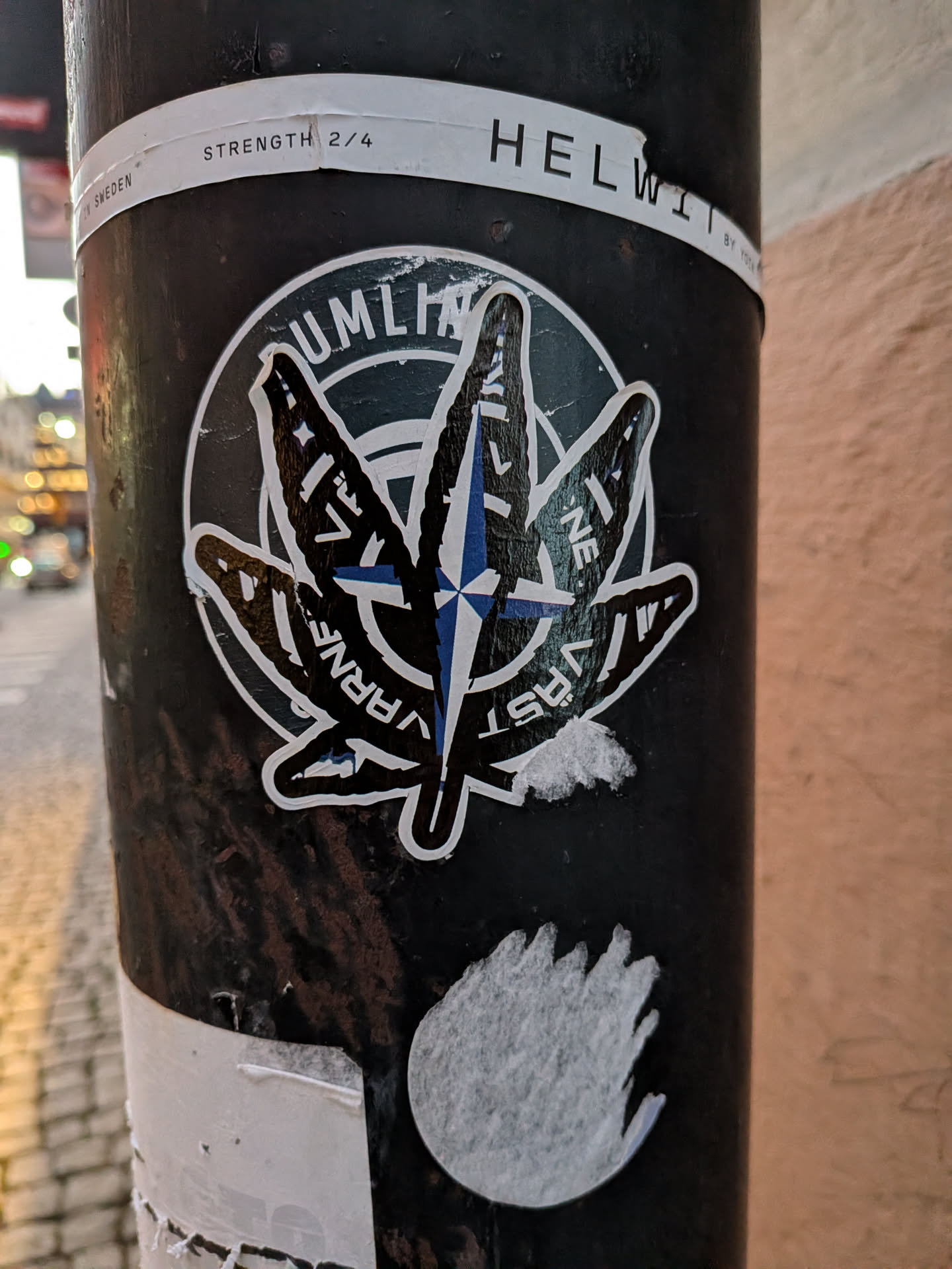 The central sticker features a stylized cannabis leaf with five distinct lobes. Overlaid on the leaf is a compass rose design, with blue and white points indicating directions. The leaf itself is predominantly black with white outlines and some internal white detailing. The background of the sticker is a dark, textured circle with white text around its perimeter. The overall impression is a bold, graphic design with a dark color palette accented by blue and white.