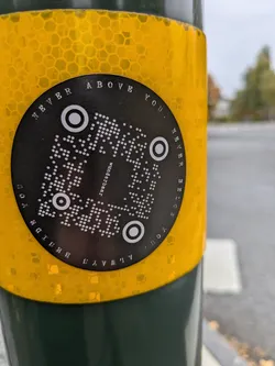 A round black and white sticker with a QR code in the center. The text "Never above you." is written around the QR code. The sticker is placed on a yellow hexagonal surface.