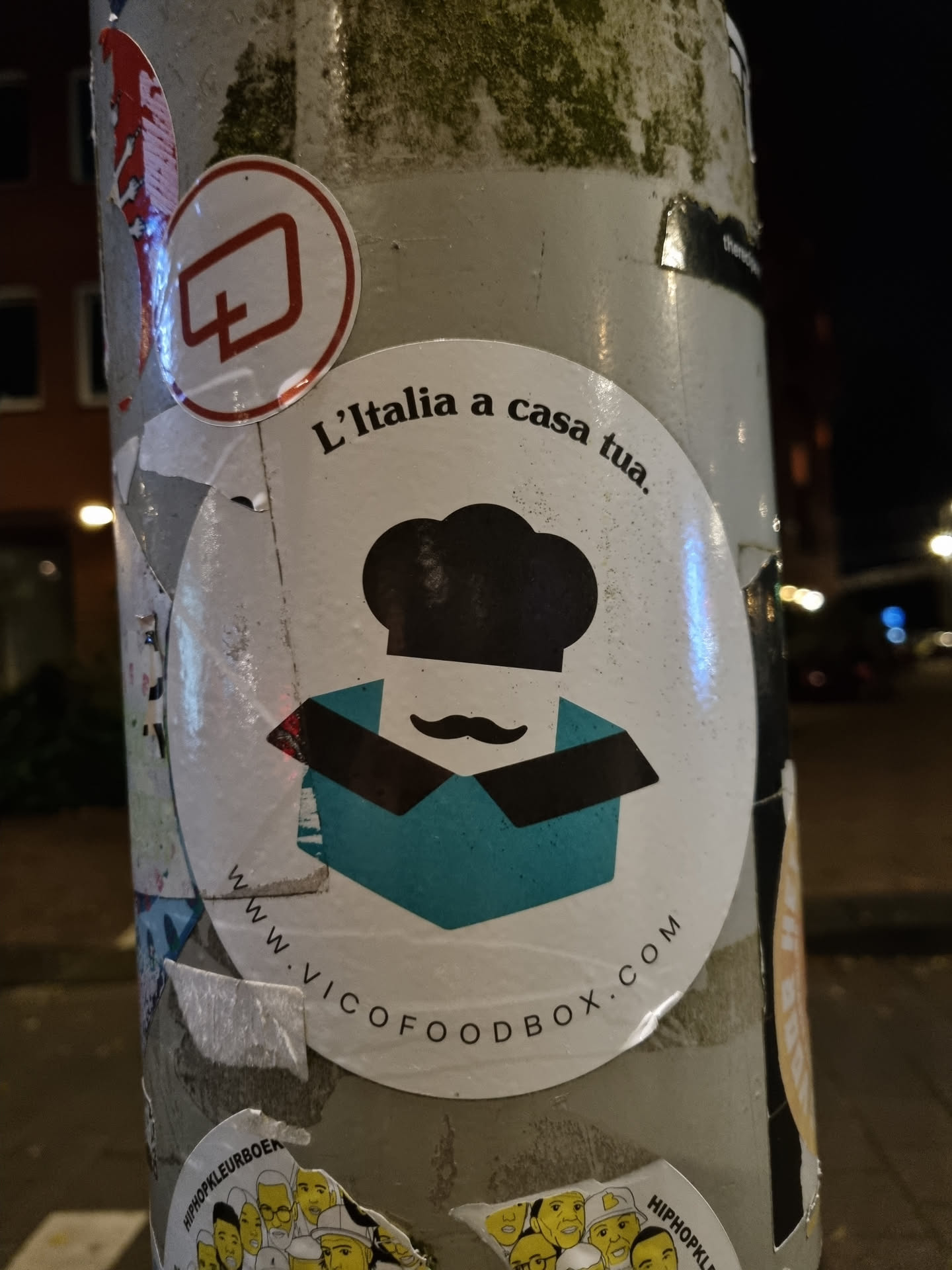 Round sticker with an illustration of a chef inside a box and the text "L'Italia a casa tua." and website address at the bottom. 