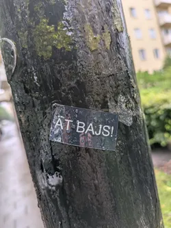 The sticker is a small, rectangular, dark-colored piece adhered to a wet, textured metal pole. It features prominent white, sans-serif capital letters on a black or very dark grey background, spelling 'ÄT BAJS!'. The surface of the sticker is visibly wet, covered in numerous small water droplets that create reflections and give it a textured appearance. The edges appear slightly worn and possibly lifted in places, typical of an outdoor sticker exposed to elements.
