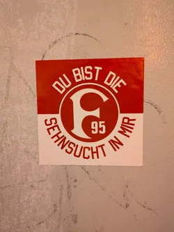 Street sticker A square football sticker divided horizontally into red and white sections. The top red section features arched white text reading 'DU BIST DIE', while the bottom white section displays arched red text reading 'SEHNSUCHT IN MIR'. In the center sits the circular crest of Fortuna Düsseldorf, showcasing the stylized white 'F' and the number '95' inside a red disc.