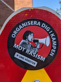 A sticker featuring a person holding a megaphone with the text "Organise yourself in the fight against racism" in Swedish. The sticker also includes the logo of Ung Vänster (Young Left), a Swedish political youth organisation.