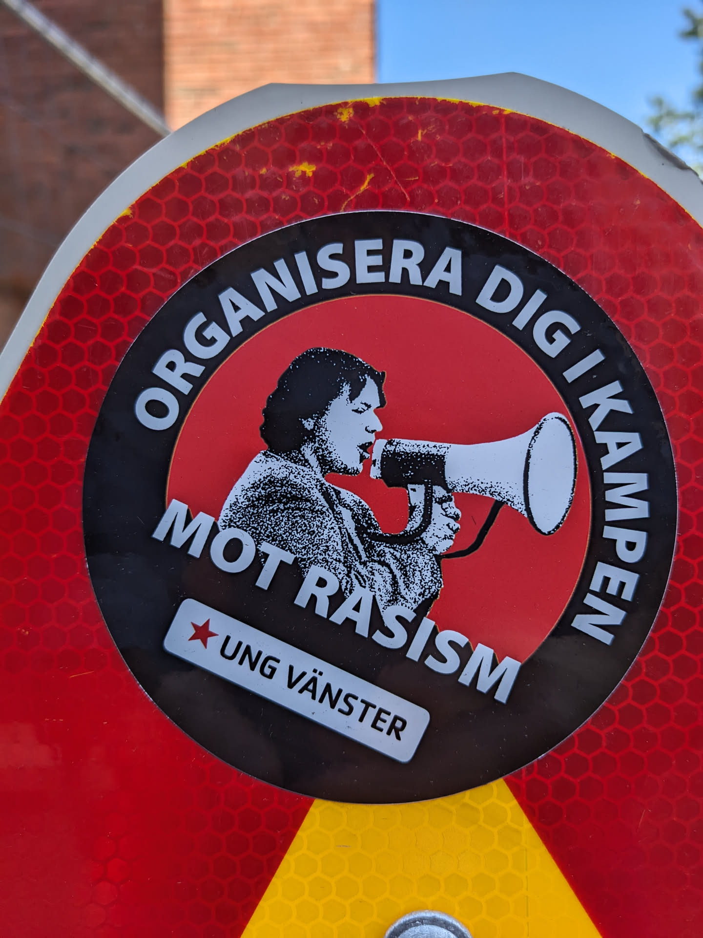 A sticker featuring a person holding a megaphone with the text "Organise yourself in the fight against racism" in Swedish. The sticker also includes the logo of Ung Vänster (Young Left), a Swedish political youth organisation.