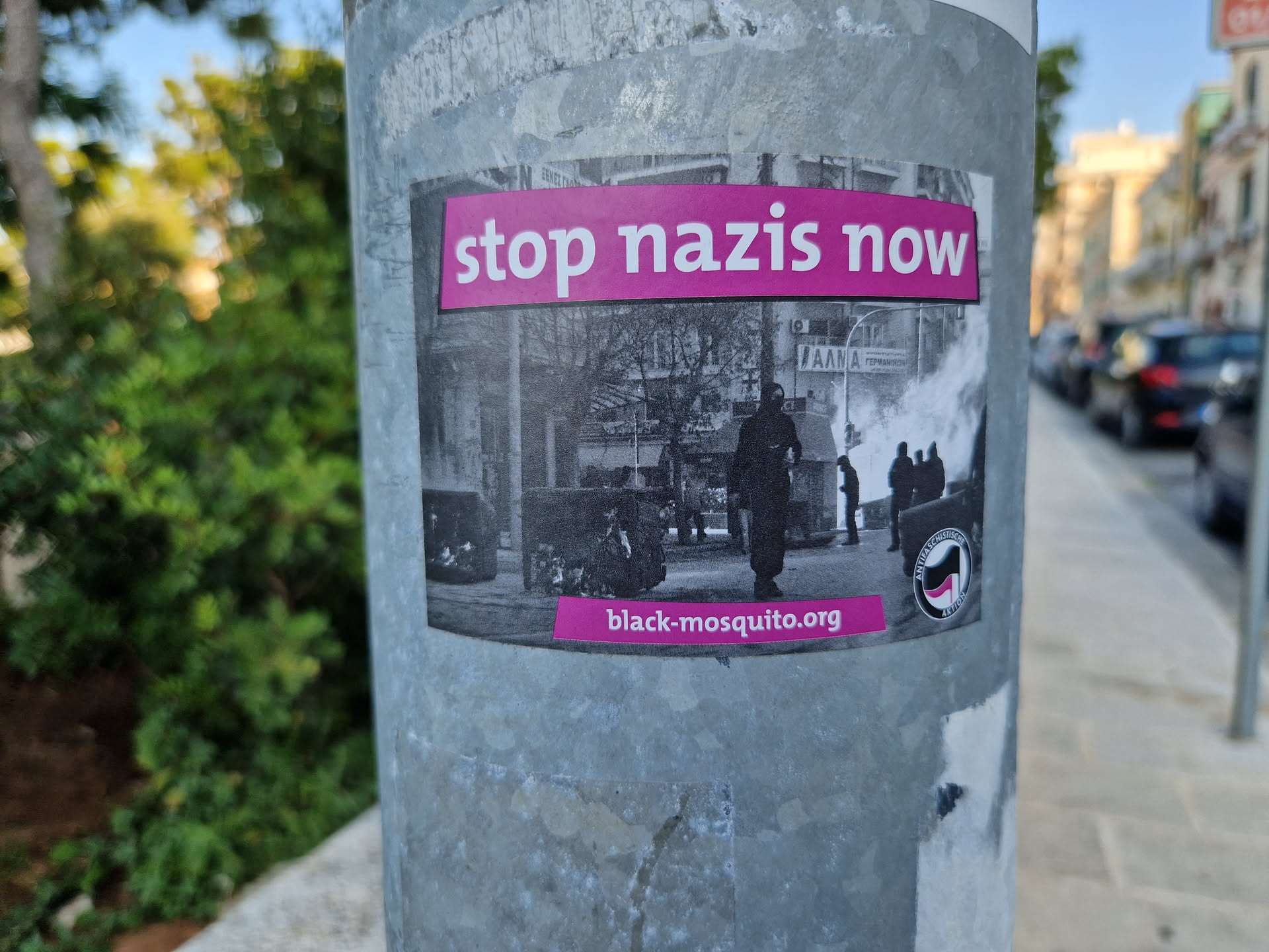 A sticker featuring a black and white image of a protest scene with the words 'Stop Nazis Now' in pink text at the top. At the bottom, the URL black-mosquito.org is visible. A logo is visible on the sticker's bottom right corner.