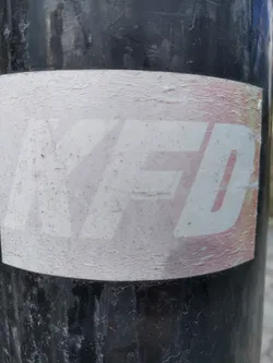 A worn sticker with the letters YFD in a stylized font. The sticker shows signs of wear and tear, suggesting it has been exposed to the elements for some time. The background is primarily white with a subtle gradient, and the lettering is slightly raised from the surface.