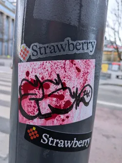 The central sticker is a rectangular piece with a white background heavily splattered with various shades of red, ranging from bright crimson to darker, almost maroon spots. The splatters are unevenly distributed, creating a dynamic and somewhat chaotic texture. Overlaid on this red-splattered canvas is a prominent, thick black graffiti tag or script. The lines of the tag are fluid, interconnected, and highly stylized, forming an illegible but energetic word or symbol. The black ink appears to be applied directly over the red splatters, giving it a layered effect. The overall appearance is raw, energetic, and embodies a street art aesthetic.