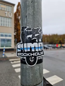 A black and white sticker with the word SUMO and the skyline of Stockholm. The sticker is slightly damaged. It's affixed to a metal pole.