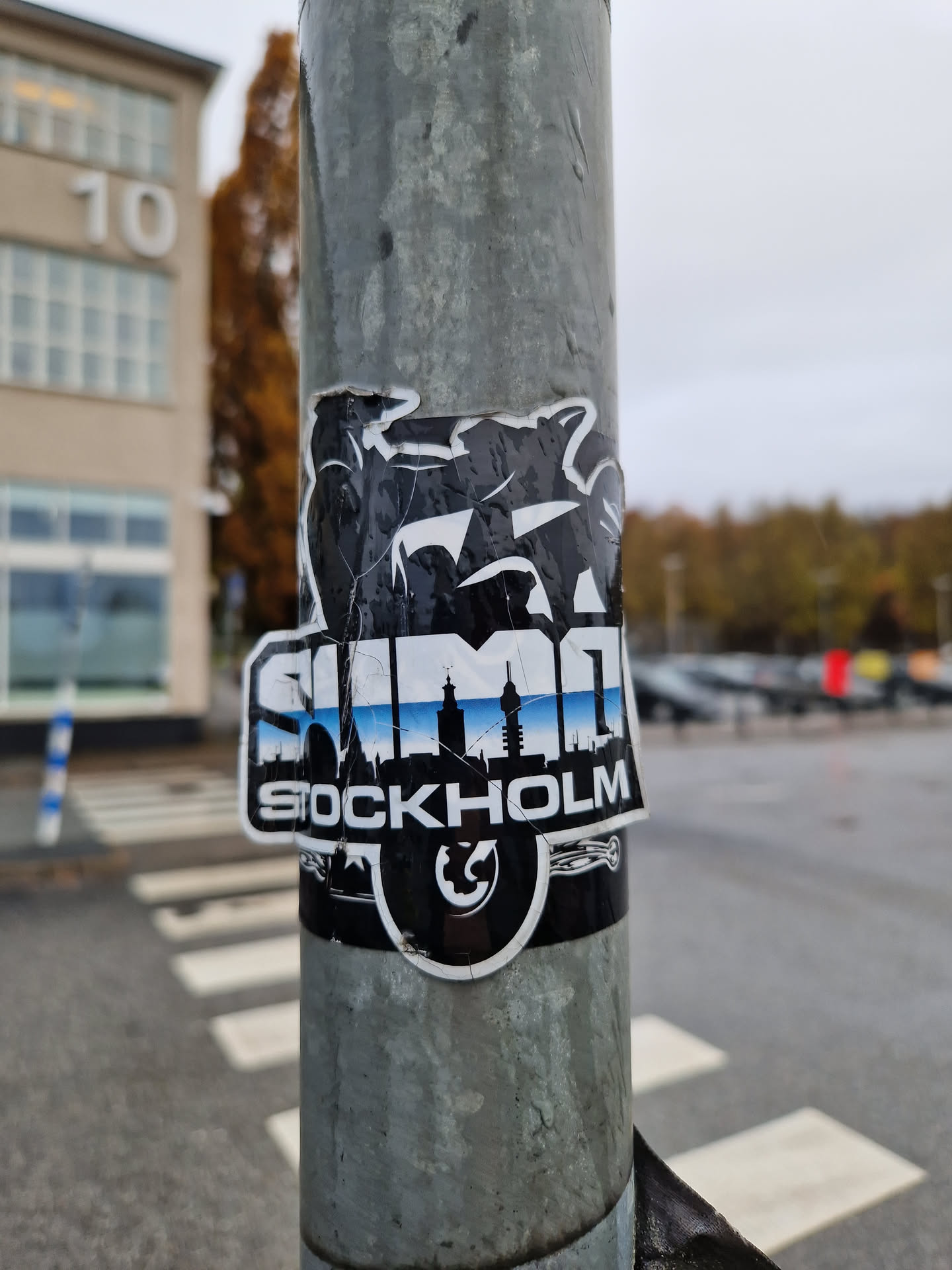 A black and white sticker with the word SUMO and the skyline of Stockholm. The sticker is slightly damaged. It's affixed to a metal pole.
