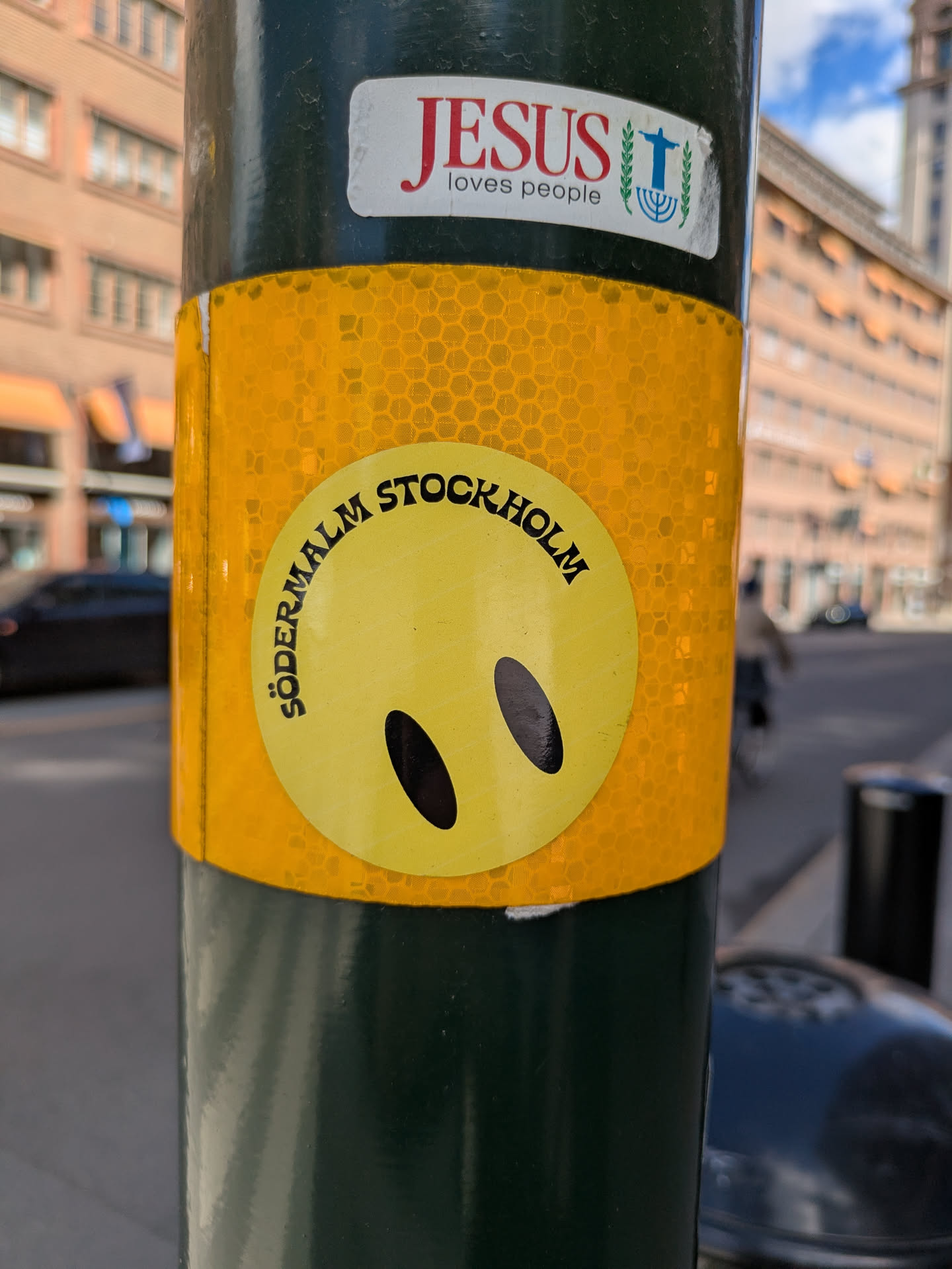 A round yellow sticker on a green pole features a smiley face with simple oval eyes and no mouth. The words "Södermalm Stockholm" are written in black around the top half of the circle. The sticker has a reflective, honeycomb-like background. Above the sticker is another sticker saying "Jesus loves people" with an image containing a cross and menorah.