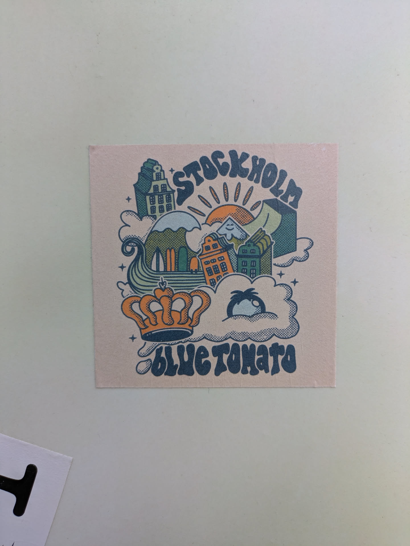 A square sticker featuring a cartoon illustration of Stockholm, Sweden. The illustration includes buildings, mountains, a sun, clouds, and a crown. The text "STOCKHOLM" is at the top and "BLUE TOMATO" at the bottom.