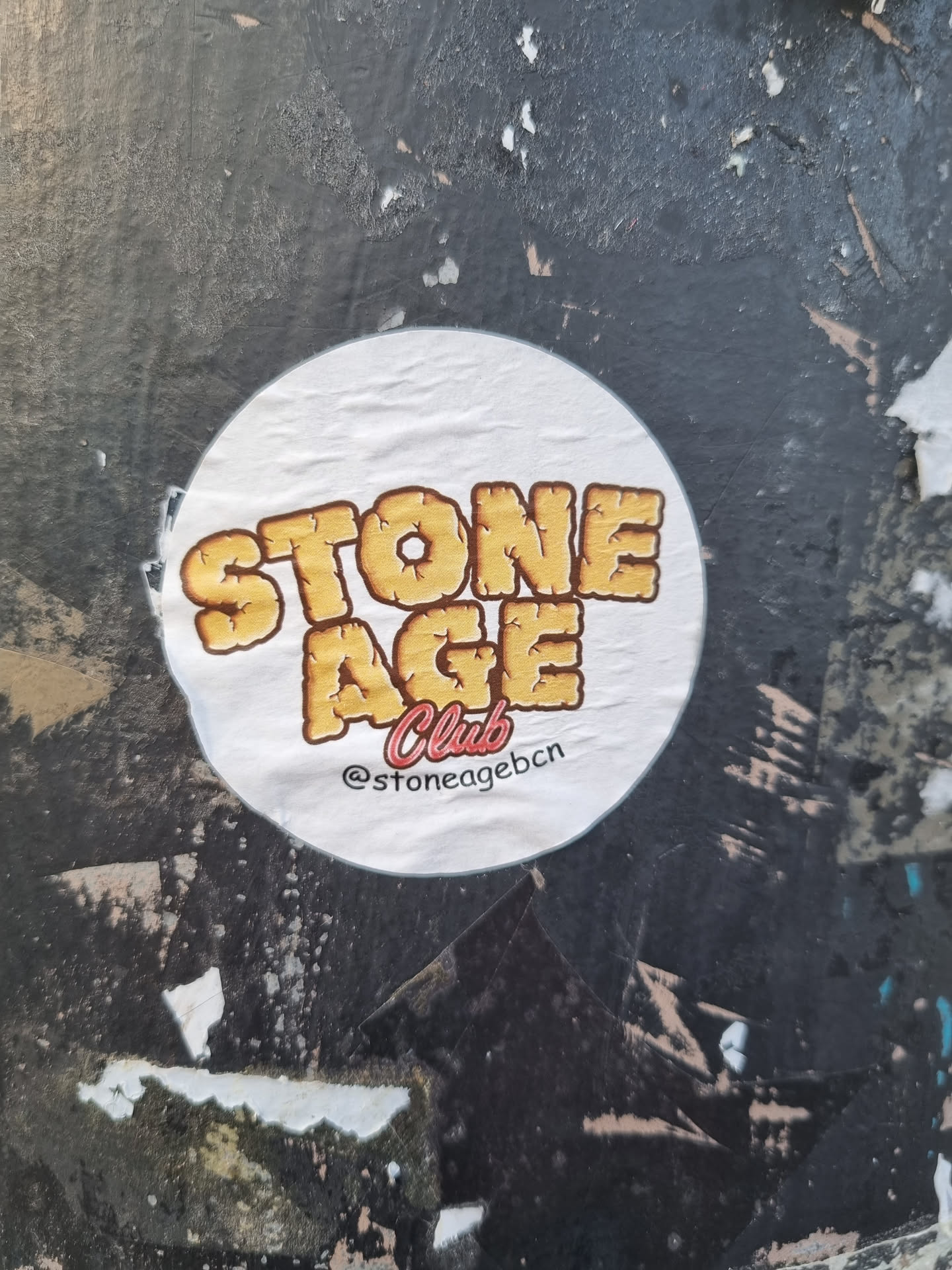 Round sticker with the text "STONE AGE Club" in a vintage style font. The text is yellow/gold with a rocky texture. The background of the sticker is white and there is a social media handle on the bottom. The sticker is affixed to a dark gray, textured surface.
