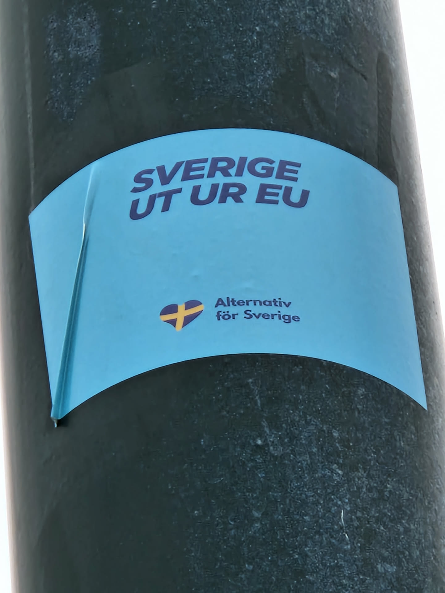 A light blue sticker affixed to a pole. It displays the text "SVERIGE UT UR EU" (Sweden out of the EU) in bold purple letters. Below, it features a stylized Swedish flag heart and the text "Alternativ för Sverige" (Alternative for Sweden).