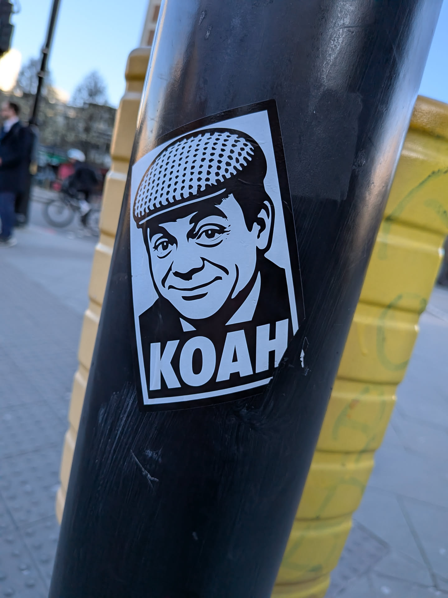 A rectangular, black and white sticker featuring a stylized, almost cartoonish, portrait of a man. He is depicted from the chest up, wearing a flat cap with a dotted texture and a collared shirt under a jacket. The man has a friendly, slightly mischievous smile and is looking slightly to the right. Below the portrait, in large, bold, white capital letters, is the word 'KOAH'. The sticker is applied to a dark pole, showing minor wear and slight peeling at the bottom right edge.