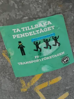 A square-shaped sticker with a vibrant teal-green background, featuring a political message conveyed through text and stick figures. The top text reads 'TA TILLBAKA PENDELTÅGET' (Take back the commuter train) in bold white letters. Below this, three black stick figures with raised arms are depicted carrying a blue, segmented object resembling a train car. To their left, a single black stick figure wearing a top hat and a yellow collar appears distressed, with hands clasped near its neck. The bottom text reads 'FRÅN TRANSPORTFÖRETAGEN' (From the transport companies). In the bottom right corner, there's a circular logo with 'ALLT ÅT ALLA' (All for All) and 'STOCKHOLM' encircling a stylized symbol, possibly an 'A' within a circle. The sticker shows signs of wear, including creases and slight discoloration, indicating it's a physical sticker adhered to a mottled grey, possibly metallic, surface with yellow paint streaks.