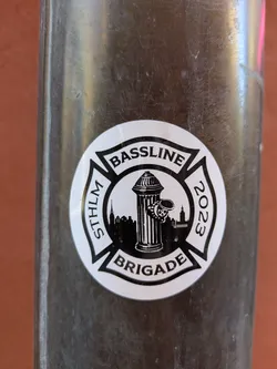 A round, black and white sticker featuring the words "BASSLINE" and "BRIGADE" with "STHLM 2023" on the sides, arranged around a fire hydrant and the Stockholm skyline in the center.
