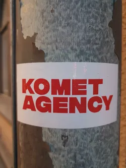 A red and white sticker with the words "KOMET AGENCY" in bold red letters on a white background. The sticker is affixed to a weathered metal pole.