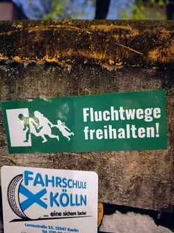 A green sticker with a pictogram and the text "Fluchtwege freihalten!" (Keep escape routes clear!) in German. Below, there is another sticker from a driving school in Cologne, Germany.