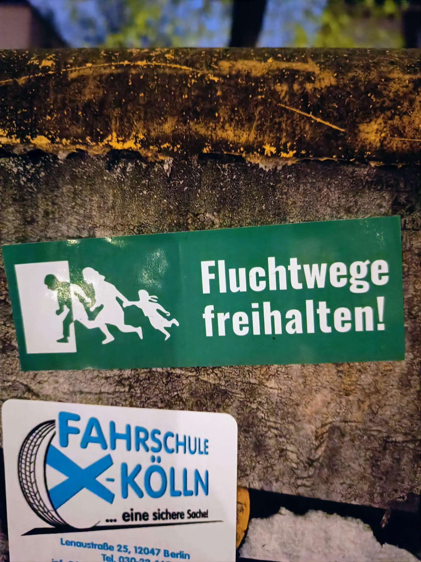 A green sticker with a pictogram and the text "Fluchtwege freihalten!" (Keep escape routes clear!) in German. Below, there is another sticker from a driving school in Cologne, Germany.