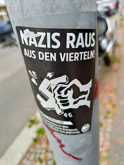 A black and white sticker with the text "Nazis raus aus den Vierteln!" (Nazis out of the neighborhoods!) and an illustration of a fist crushing a swastika.  The sticker is affixed to a metal pole.