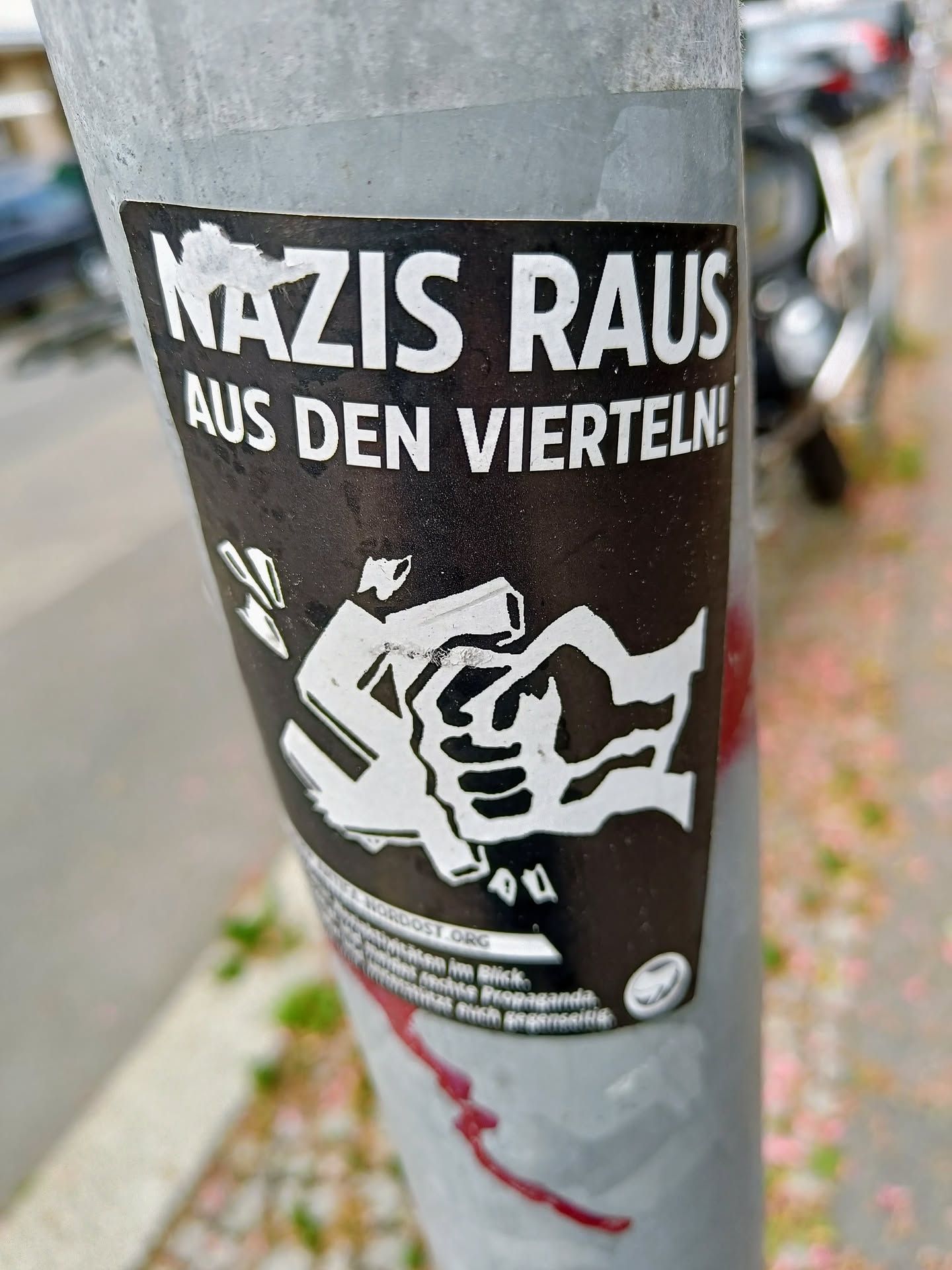 A black and white sticker with the text "Nazis raus aus den Vierteln!" (Nazis out of the neighborhoods!) and an illustration of a fist crushing a swastika.  The sticker is affixed to a metal pole.