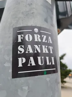 Black and white sticker with the text "Forza Sankt Pauli" and a skull symbol.  The sticker is affixed to a grey metal pole.