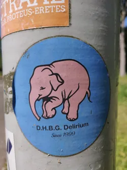 A round sticker on a pole with a light blue background featuring a pink elephant. The text below the elephant reads: "D.H.B.G. Delirium Since 1999"