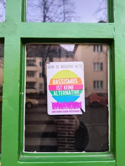 A sticker affixed to a window pane, proclaiming 'Rassismus ist keine Alternative' in bold letters, encircled by a rainbow design. Below, an illustration depicts a multitude of figures. At the bottom, smaller print reads 'Aufstehen gegen Rassismus'. The overall message is a strong condemnation of racism.