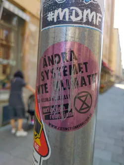 A pink round sticker on a metal pole with the text "Ändra systemet inte klimatet" (Change the system, not the climate) and the Extinction Rebellion logo.  The website www.extinctionrebellion.se is also visible.