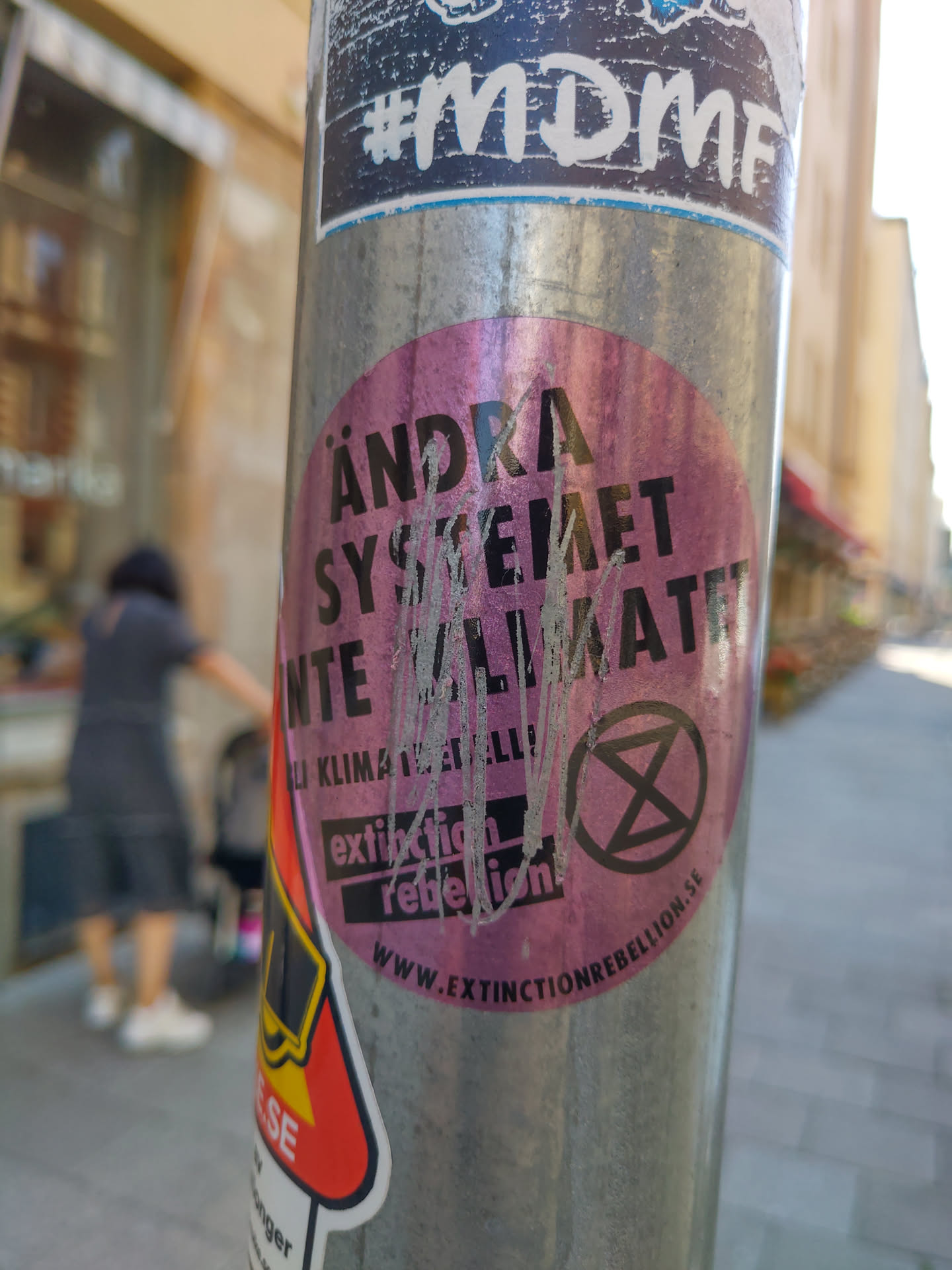 A pink round sticker on a metal pole with the text "Ändra systemet inte klimatet" (Change the system, not the climate) and the Extinction Rebellion logo.  The website www.extinctionrebellion.se is also visible.