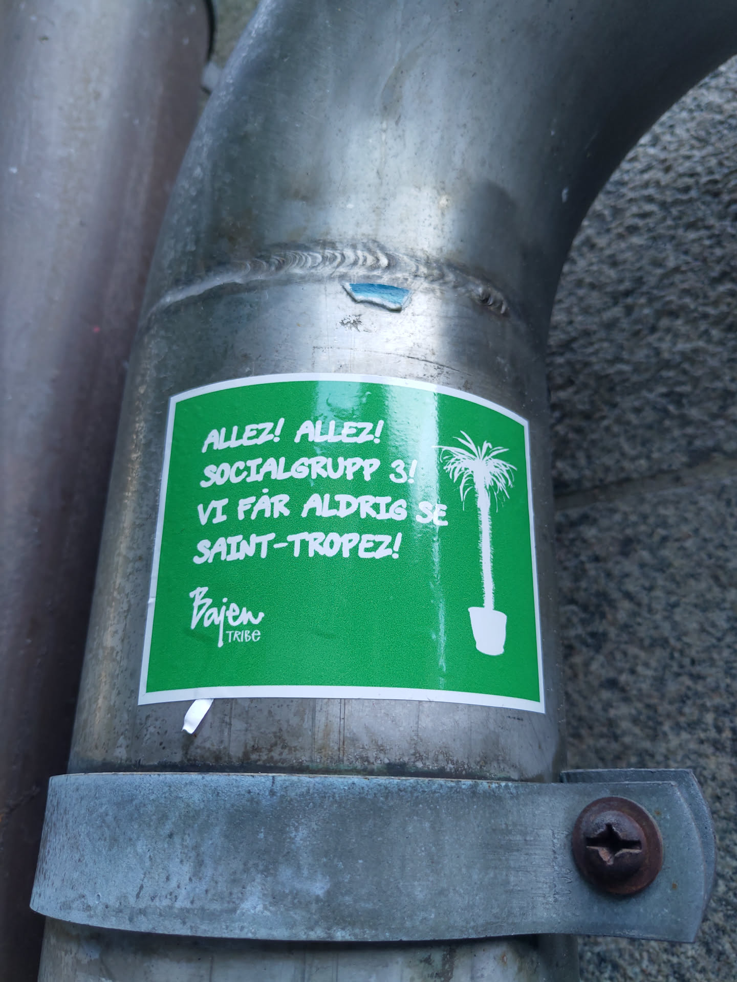 A green sticker with white text and a drawing of a plant in a pot. The text says: Allez! Allez! Socialgrupp 3! Vi får aldrig se Saint-Tropez! There is also a logo that says Pajen Tribe.
