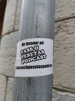 A sticker for the PASS & PESETAS PODCAST. The sticker is white with black text. It's attached to a metal pole.