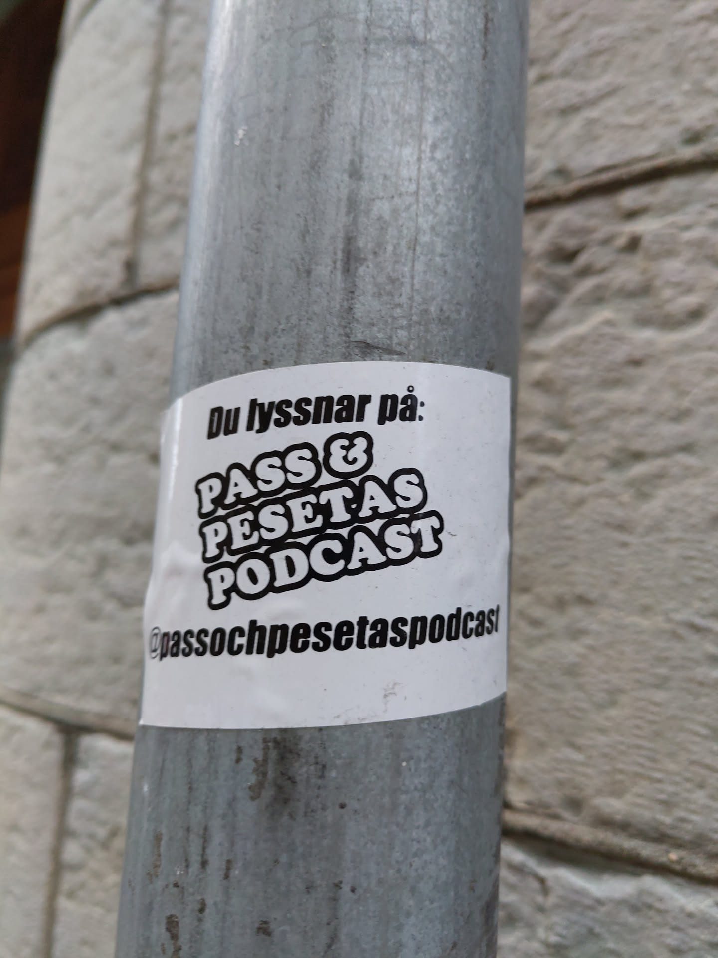 A sticker for the PASS & PESETAS PODCAST. The sticker is white with black text. It's attached to a metal pole.