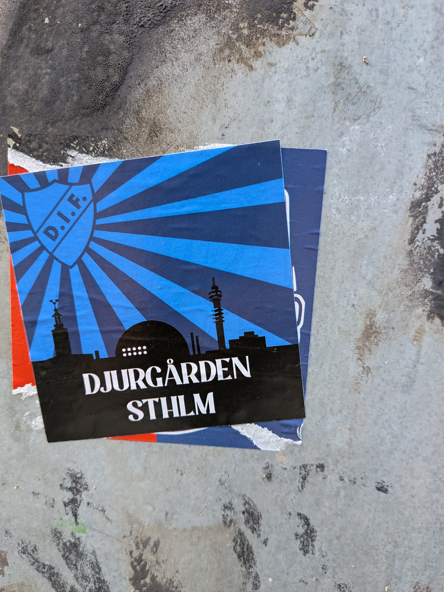 The sticker features a blue shield with 'D.I.F.' inside it, set against a background of radiating blue lines. Below the shield is a black silhouette of the Stockholm skyline, including recognizable buildings like the Kaknäs Tower, and a dome-shaped structure with circular windows. The text 'DJURGÅRDEN' is prominently displayed in white capital letters, followed by 'STHLM' in slightly smaller white capital letters. The sticker appears to be slightly crumpled and is layered over other torn pieces of paper.