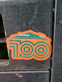 Orange and green sticker commemorating 100 years of Eventyr Kiosken. The design features the number 100 with concentric circles and a small illustration of a kiosk at the top.
