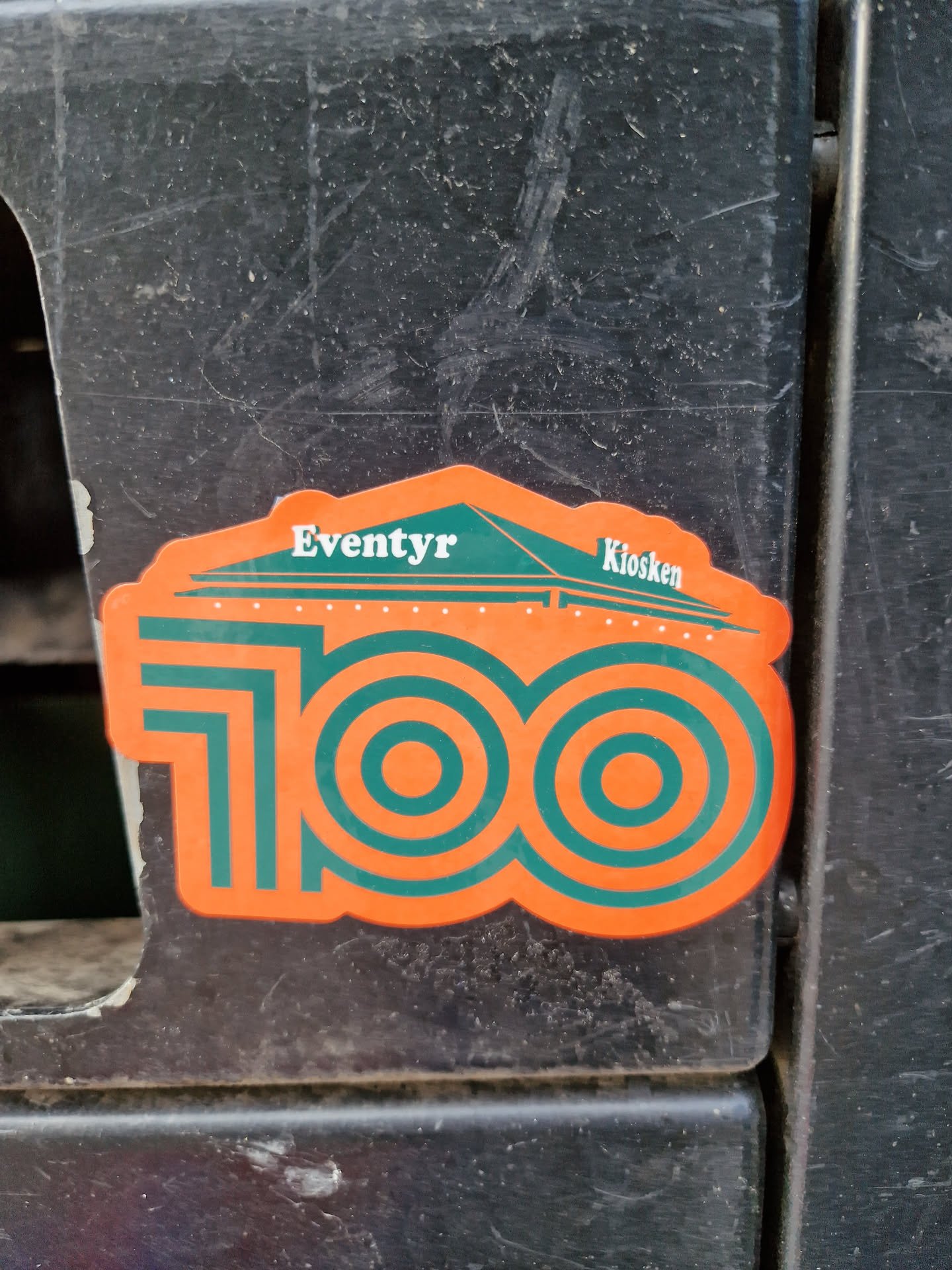Orange and green sticker commemorating 100 years of Eventyr Kiosken. The design features the number 100 with concentric circles and a small illustration of a kiosk at the top.