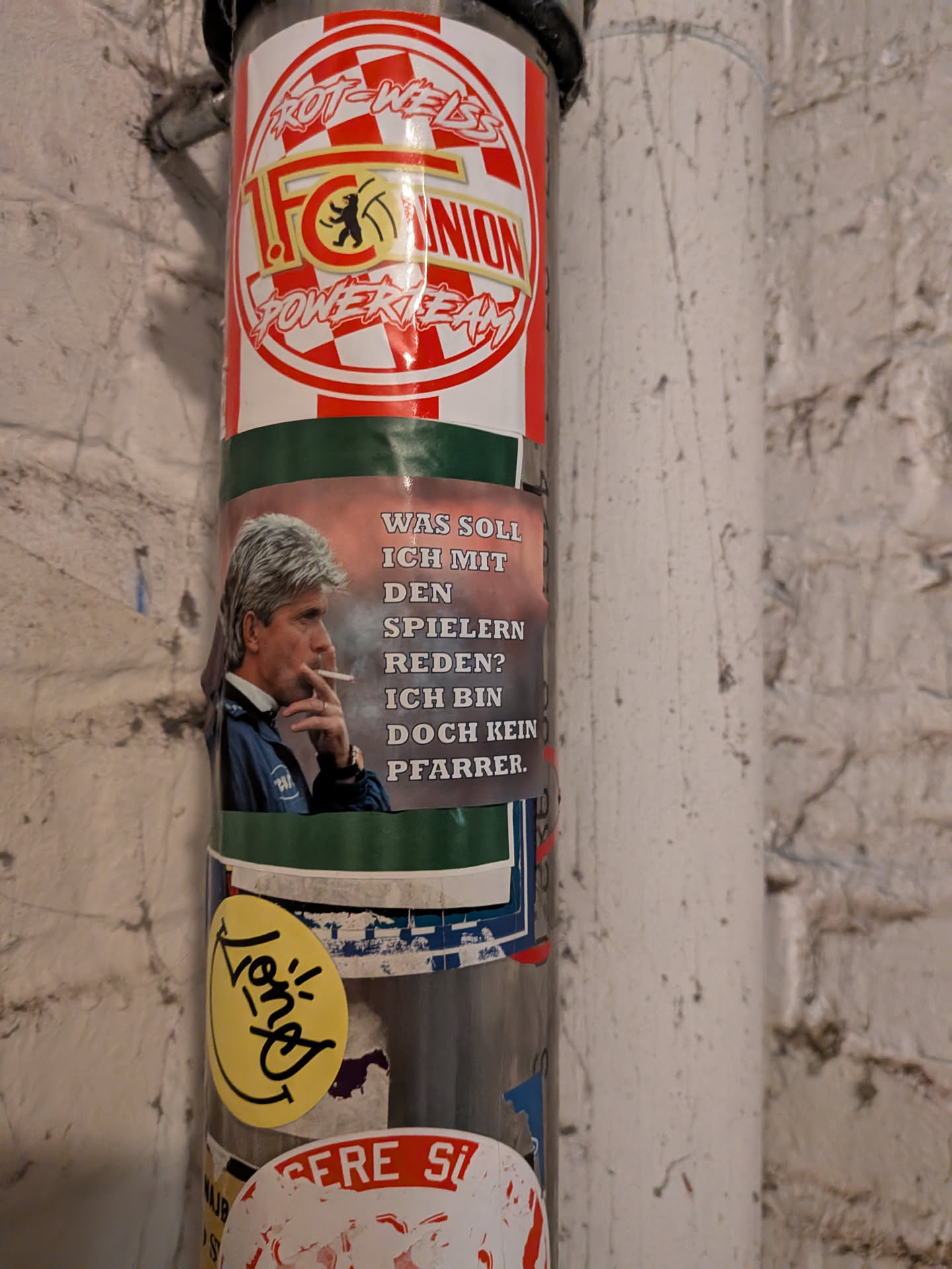 A rectangular sticker placed horizontally around a vertical pipe. The left side features a color photograph of football coach Ernst Happel in profile, holding a lit cigarette to his lips and wearing a blue training jacket. The right side features a muted, smoky background with bold, all-caps German text with drop shadows that reads: 'WAS SOLL ICH MIT DEN SPIELERN REDEN? ICH BIN DOCH KEIN PFARRER.'