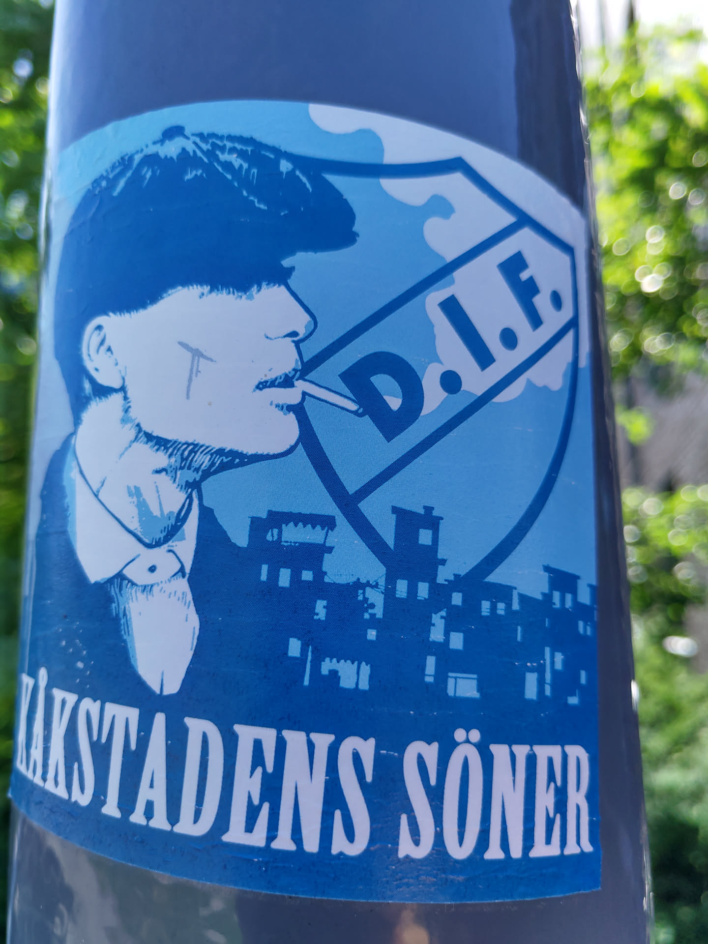 A blue and white sticker featuring a stylized illustration of a man resembling Tommy Shelby from the Peaky Blinders TV series.  He is smoking a cigarette and is set against a backdrop of buildings, with a prominent shield-shaped logo reading "D.I.F." The bottom of the sticker displays the text "ÄKSTADENS SÖNER" in white lettering.
