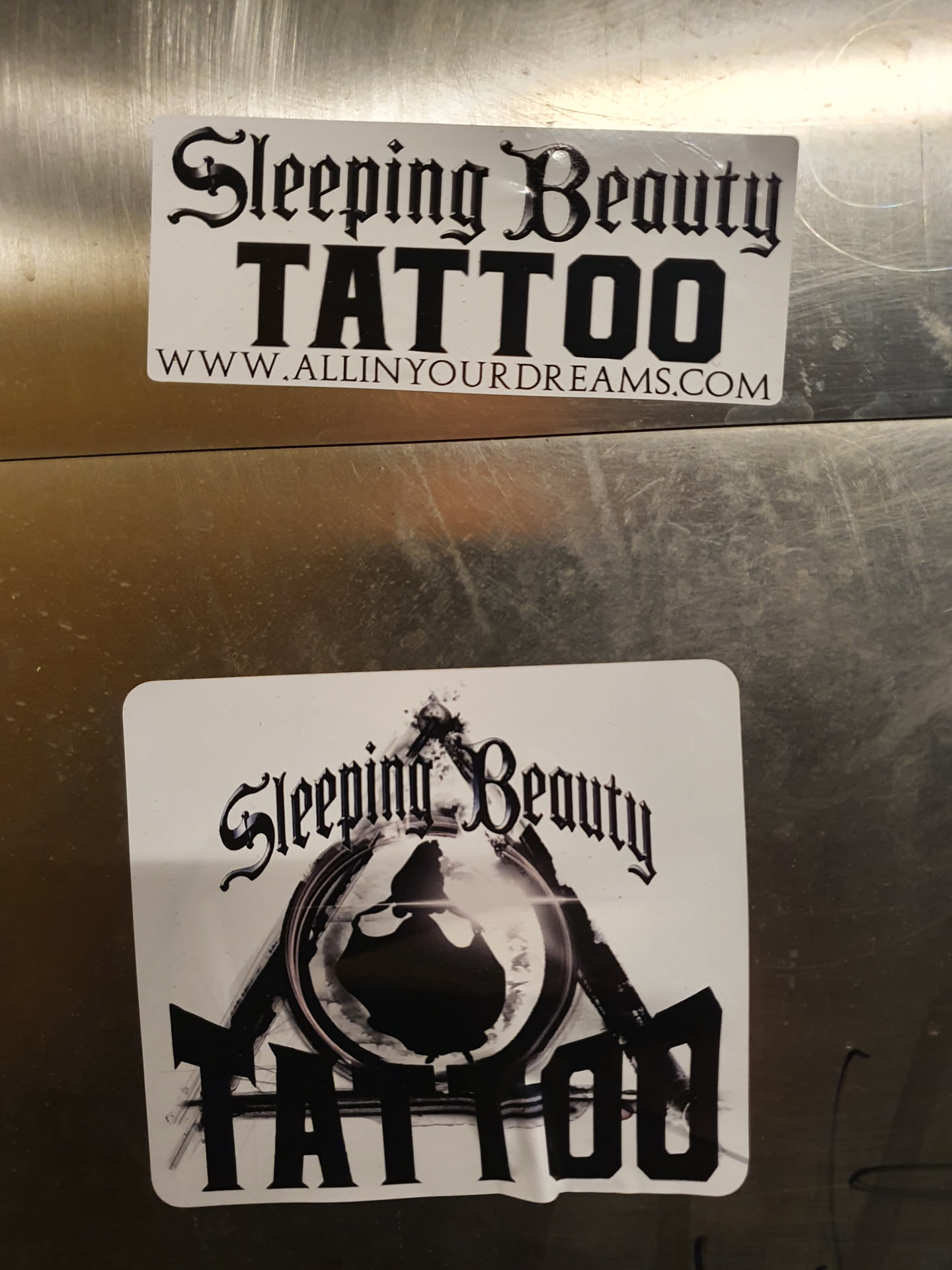 Sleeping Beauty Tattoo shop sticker. The sticker features a stylized image of Sleeping Beauty within a triangle, with the text 'Sleeping Beauty TATTOO' in a gothic-style font.  A website address is also included on a second sticker.