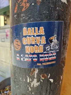 A sticker depicting a cyclist riding a bike with a dark blue background and orange text. The text reads "DALLA CURVA NORD A CAPO NORD" and includes the logo of Soc. Valdinievole.